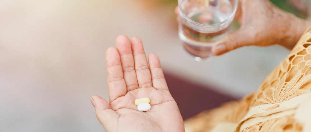 Kobieta trzyma na dłoni kilka suplementów w tabletkach oraz szklankę wody
