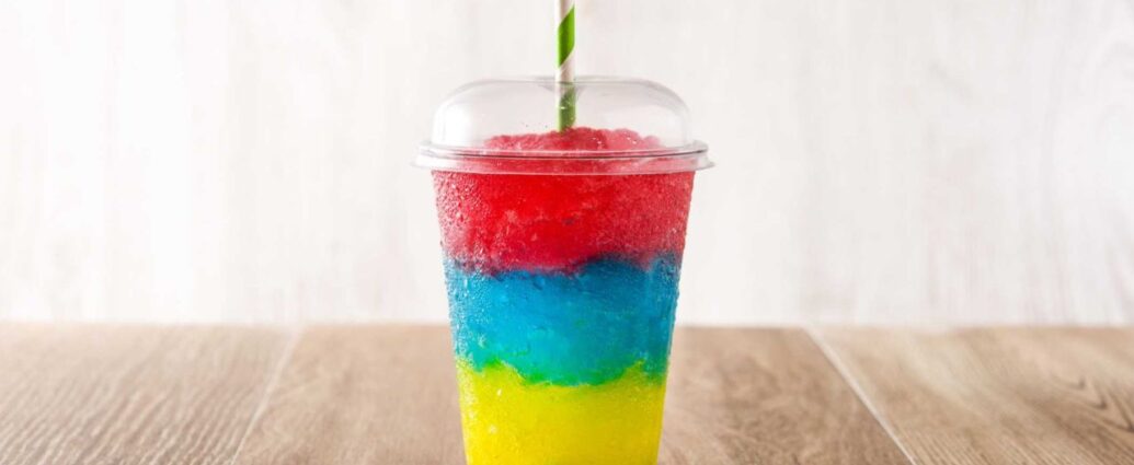 Trójkolorowa granita w przezroczystym kubku z kopułką, ustawiona na drewnianym blacie. Widoczne warstwy w intensywnych kolorach: żółtym, niebieskim i czerwonym, podkreślają wakacyjny charakter napoju i różnorodność smaków popularnych latem w punktach sezonowych. Więcej informacji o najchętniej wybieranych smakach granity można znaleźć na stronie z artykułem