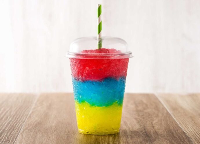 Trójkolorowa granita w przezroczystym kubku z kopułką, ustawiona na drewnianym blacie. Widoczne warstwy w intensywnych kolorach: żółtym, niebieskim i czerwonym, podkreślają wakacyjny charakter napoju i różnorodność smaków popularnych latem w punktach sezonowych. Więcej informacji o najchętniej wybieranych smakach granity można znaleźć na stronie z artykułem